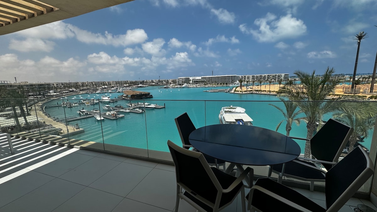 A spacious balcony features a round table and black lounge chairs, offering an unobstructed view of the marina. The turquoise waters reflect the sky, while boats are anchored in the distance. Palm trees add a touch of greenery to the coastal setting.