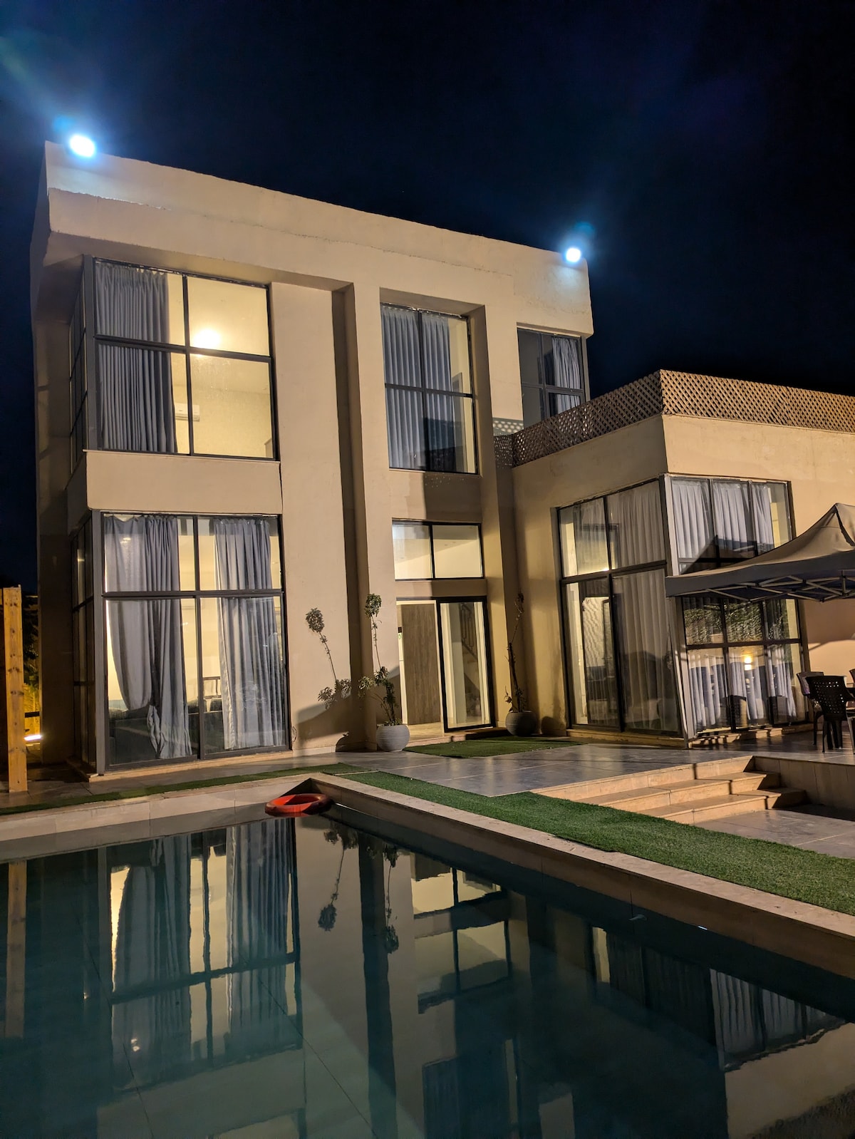 The villa's modern architecture is highlighted at night, showcasing large glass windows that reflect the soft glow of exterior lighting. A private pool lies in the foreground, complementing a spacious outdoor seating area with a canopy, creating a serene and inviting environment.