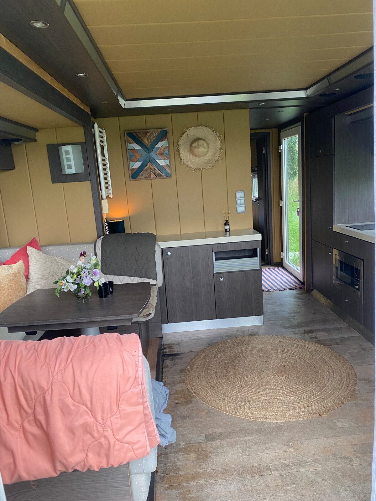 An inviting interior space is showcased, featuring a comfortable seating area with decorative cushions and a small table. Natural light fills the room through a door leading outside, enhancing the earthy tones of the wood paneling and the round rug on the floor.