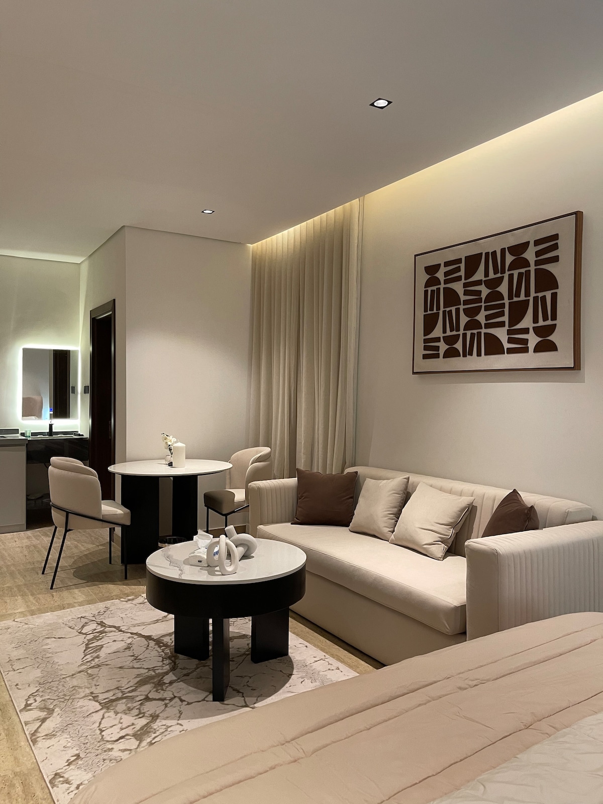 A modern living space features a comfortable sofa against a softly lit wall. A round coffee table is positioned in front of the sofa, accompanied by two chairs. Light colors and minimalist decor create a serene ambiance, with an art piece mounted above the sofa.