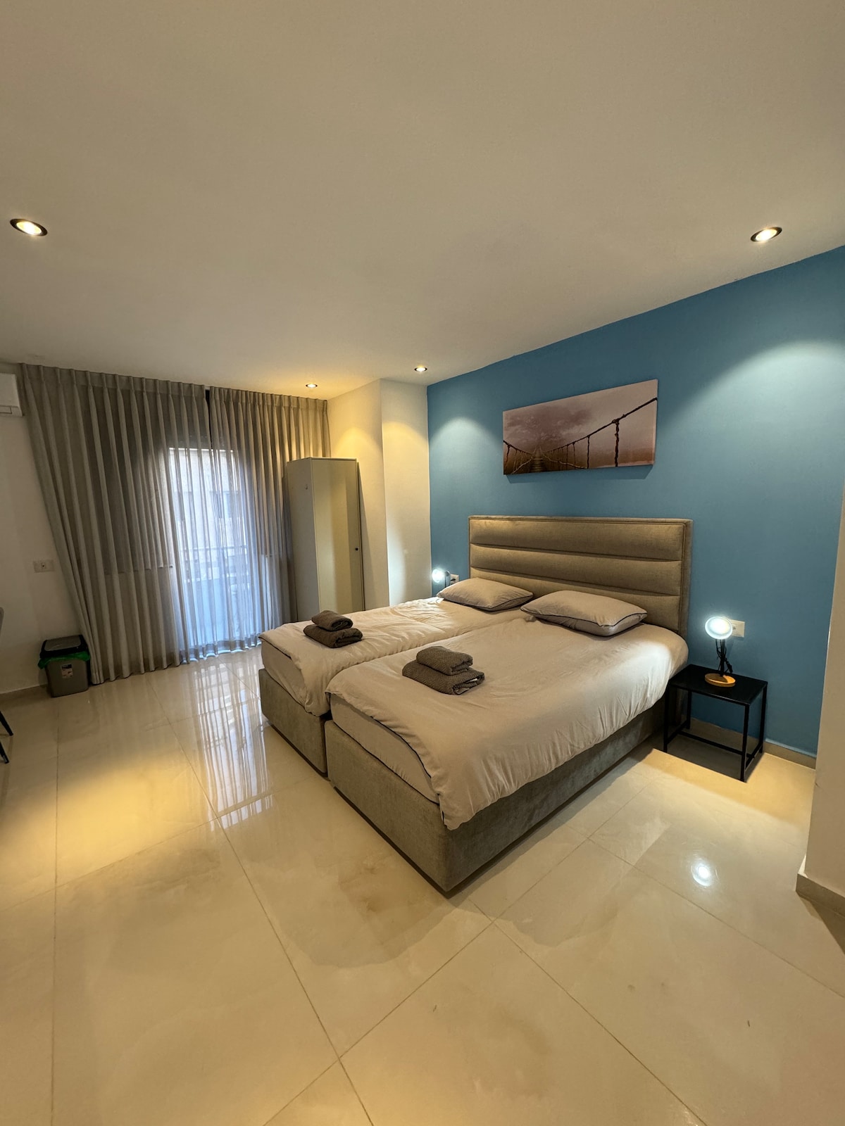The sleeping area features two spacious beds arranged side by side, each topped with neatly folded towels. Soft lighting is provided by bedside lamps on either side. A large window is dressed with sheer curtains, allowing natural light to brighten the room, which is adorned with a calming blue accent wall.