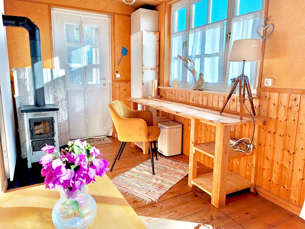 A light-filled interior space features a wooden desk positioned beside a large window, offering a view of the outdoors. A cozy chair sits at the desk, while a small wood stove provides warmth. Fresh flowers are displayed in a clear vase on the table, enhancing the natural ambiance.