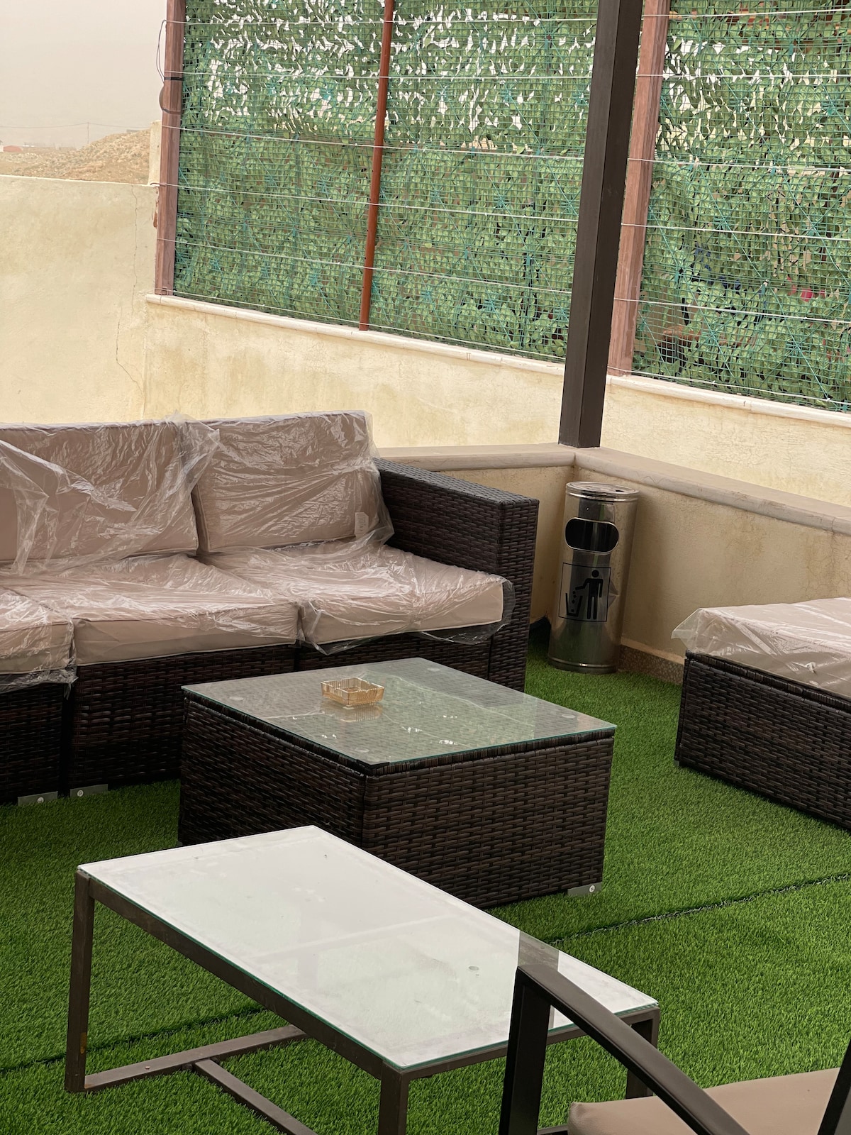 An outdoor seating area features a set of dark wicker furniture, including a couch and a pair of chairs, covered in protective plastic. A glass-topped coffee table sits at the center on a grassy green artificial turf, contributing to a relaxing atmosphere.