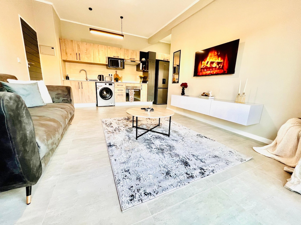 A modern living space features a plush sofa and a decorative area rug in muted tones. A sleek coffee table sits centrally, while a wall-mounted television displays an image of a cozy fireplace. The kitchen area includes essential appliances, enhancing the apartment's functionality.
