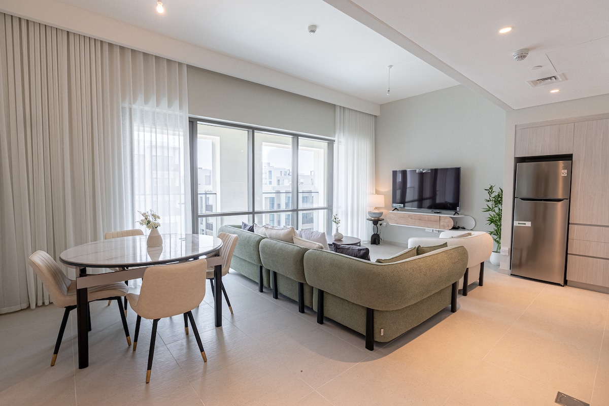 A spacious living area features a large sectional sofa arranged around a coffee table. A stylish dining table with light-colored chairs sits nearby. Floor-to-ceiling windows provide ample natural light, offering views of the outside. A modern television is mounted on the wall, and a refrigerator is visible in the corner.