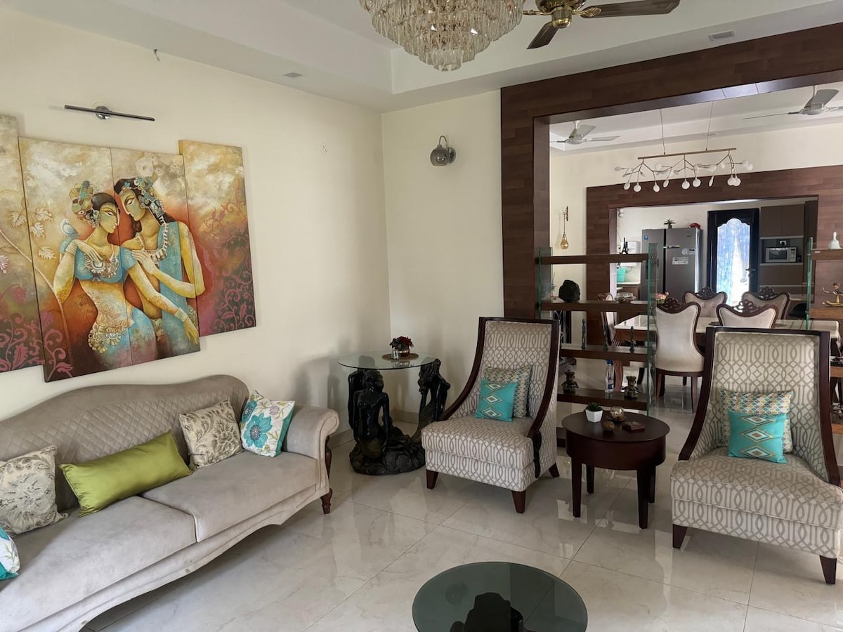 A spacious drawing room features a soft sofa adorned with colorful cushions. Two elegant chairs with intricate patterns flank a glass coffee table. A vibrant artwork depicting three figures is displayed prominently on the wall, adding character to the room.