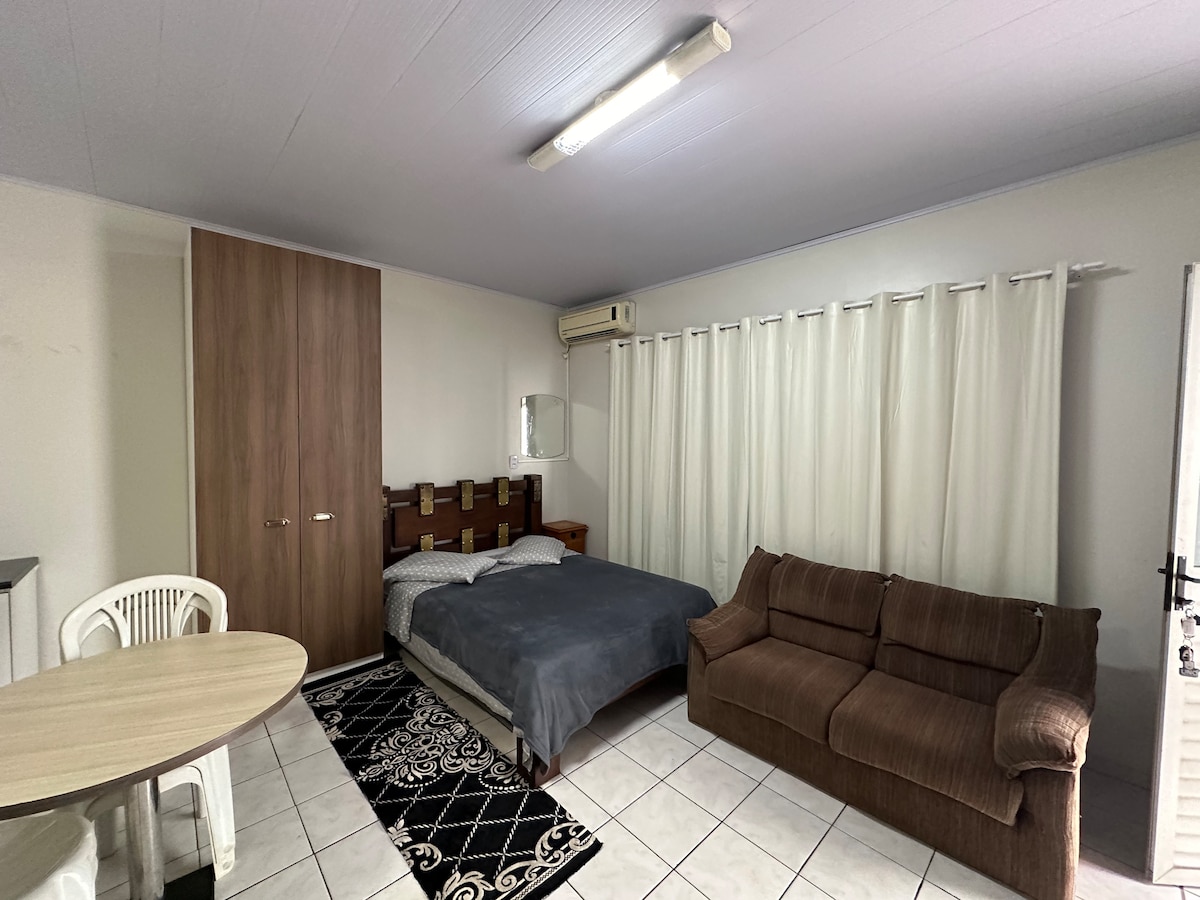 A furnished studio space features a spacious bed with grey linens, complemented by a dark sofa. A round dining table with white chairs is positioned nearby. Natural light filters through long, light-colored curtains, while a mirrored wall adds depth to the room.