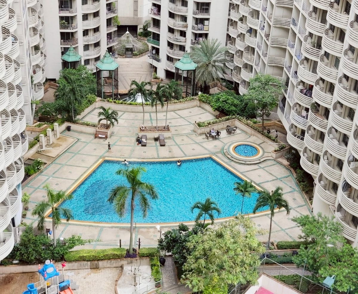 An aerial view of a spacious swimming pool, surrounded by palm trees and well-maintained gardens. The pool area features lounge chairs and shaded seating options. Buildings encircle the pool, enhancing accessibility to the communal space. The overall atmosphere is relaxed and inviting.