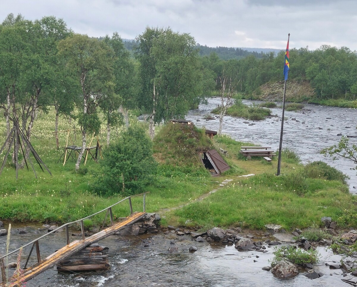 A wooden bridge crosses a flowing river, leading to an area with grassy terrain. Several benches are positioned nearby, along with a flagpole displaying a multicolored flag. The natural surroundings include trees and low shrubs, contributing to a serene outdoor environment.