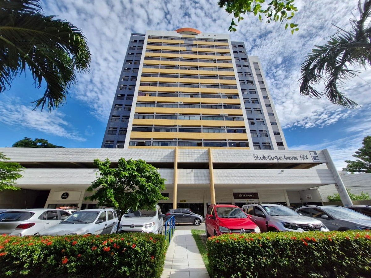 A modern high-rise building stands tall against a clear blue sky, showcasing a prominent orange rooftop feature. The ground level includes a parking area with several vehicles, landscaped greenery, and flowering plants lining the pathway leading to the entrance.