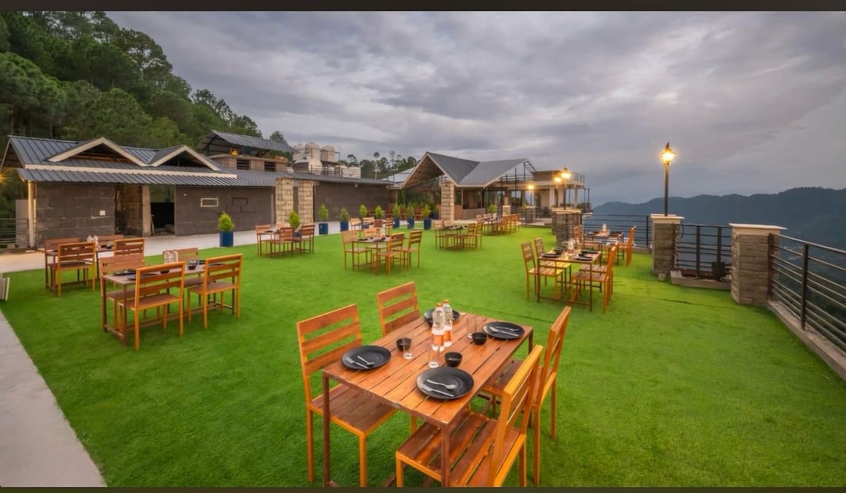 An outdoor dining area is set on a lush green lawn, furnished with wooden tables and chairs. Each table is arranged with black plates and silverware. Scenic views of the surrounding mountains can be seen in the background, under a partially cloudy sky.