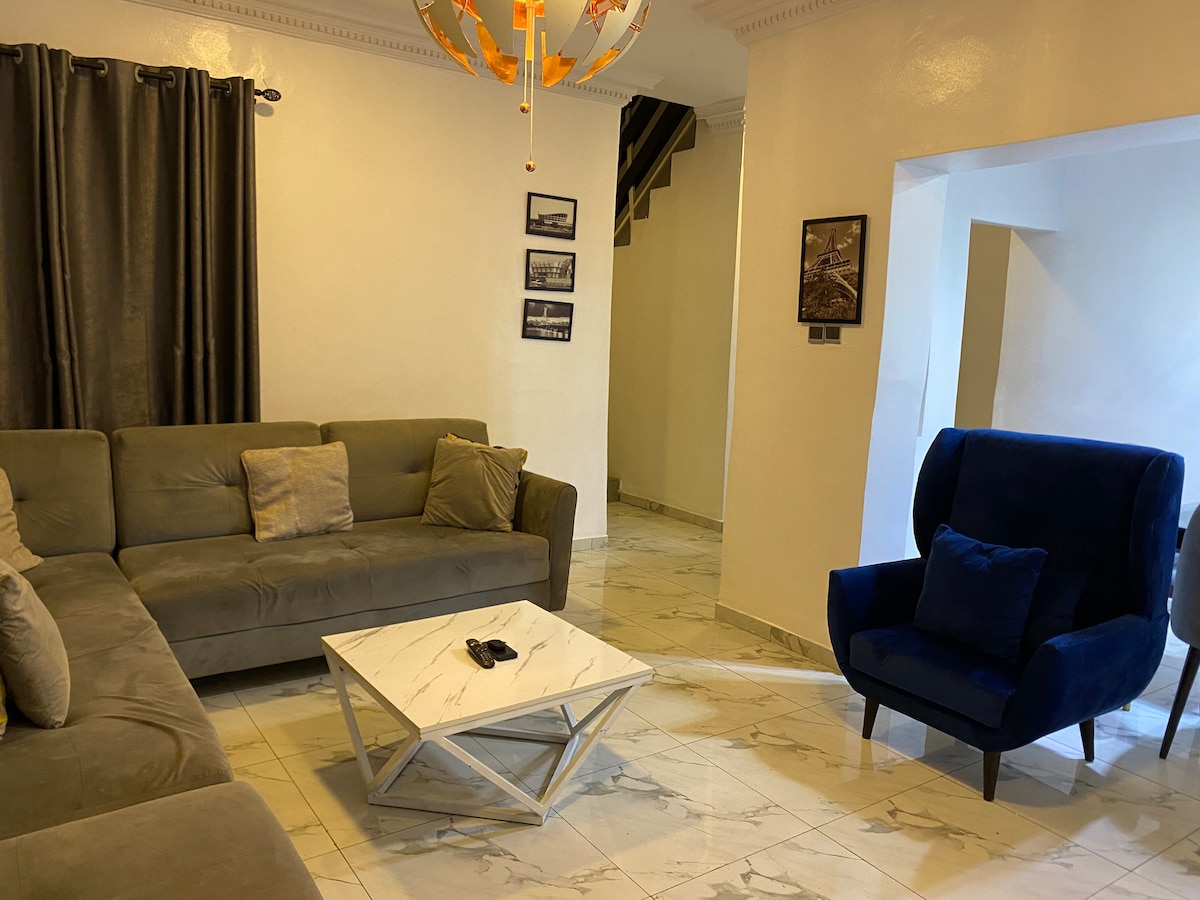 A bright living area features a large, L-shaped sectional sofa in a neutral tone. A marble coffee table sits in the center, complemented by a single blue armchair. Stylish light fixtures hang from the ceiling, while artwork adorns the walls, enhancing the overall charm.