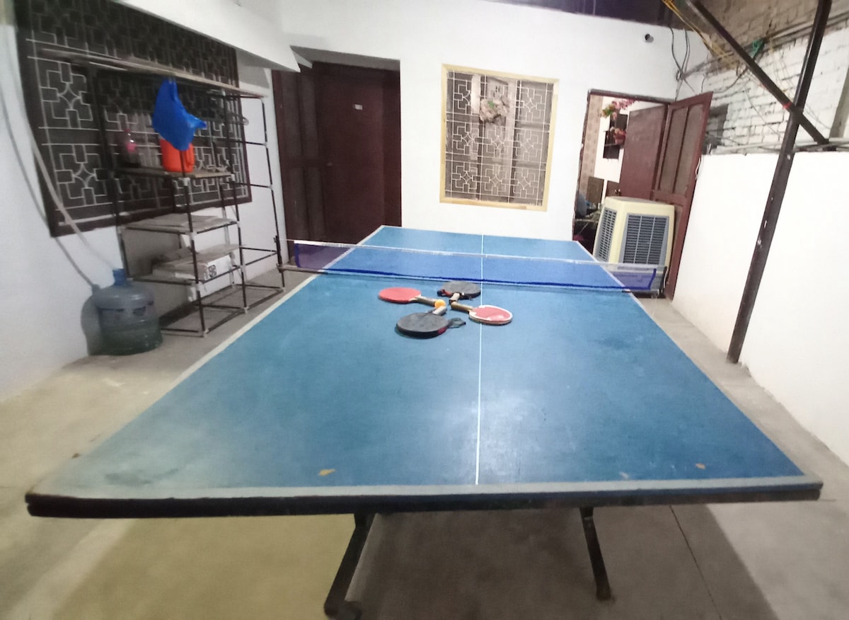 A table tennis table is prominently displayed in a spacious recreational area, surrounded by light-colored walls. Nearby, a blue paddle and a ball rest on the table surface, with an air conditioning unit visible in the background.