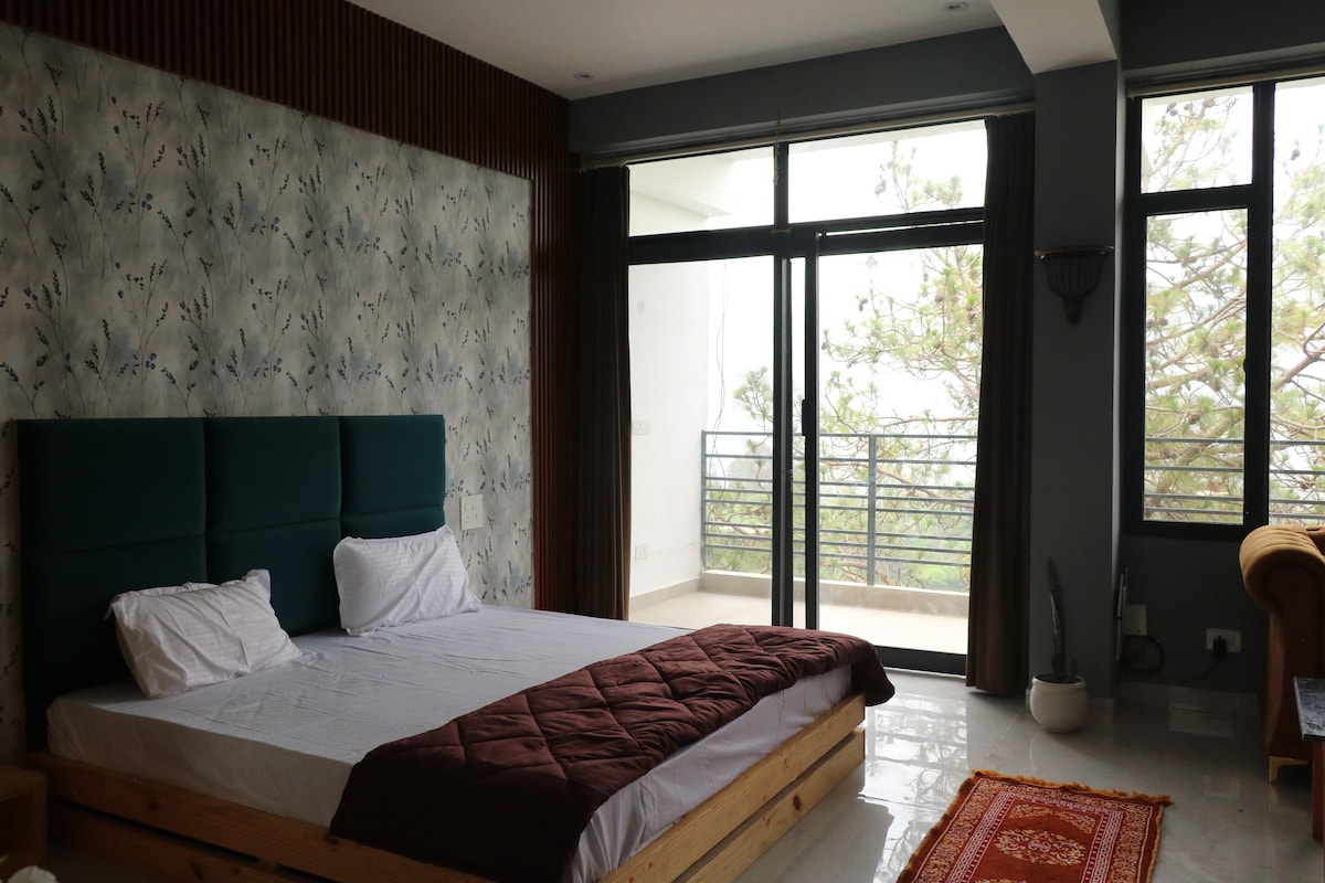 A spacious bedroom features a large bed with a dark red blanket and two white pillows. Floor-to-ceiling windows allow natural light to fill the room, framed by sleek, dark curtains. A patterned accent wall adds visual interest, complemented by a cozy seating area and a small plant.