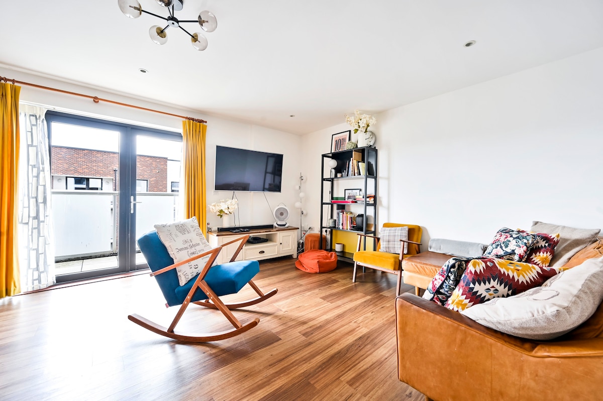 A bright living room features wooden flooring and large glass doors that open to a balcony. A flat-screen TV is mounted on a white wall opposite a mixed seating arrangement, including a rocking chair, a vibrant orange pouf, and a cozy couch adorned with colorful cushions.
