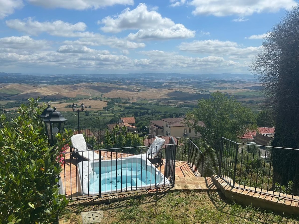 A private hot tub is positioned on a terrace, offering panoramic views of rolling hills and valleys. Lush greenery is present around the patio area, with a couple of lounge chairs arranged nearby. The sky is bright with scattered clouds, enhancing the serene landscape.
