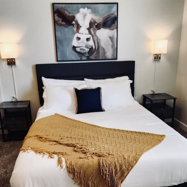 Დახვეწილი საძინებელი სუფთა თეთრი თეთრეულით, მყუდრო საბნითა და მხიარული ძროხის ხელოვნების ნიმუშებით. Წარმოდგენილია ორმაგი ტუმბოები კედელზე მიმაგრებული რბილი განათებით, პლუშის საწოლითა და ნეიტრალური ტონებით დასასვენებლად, თანამედროვე ფერმერული საცხოვრებლისთვის.