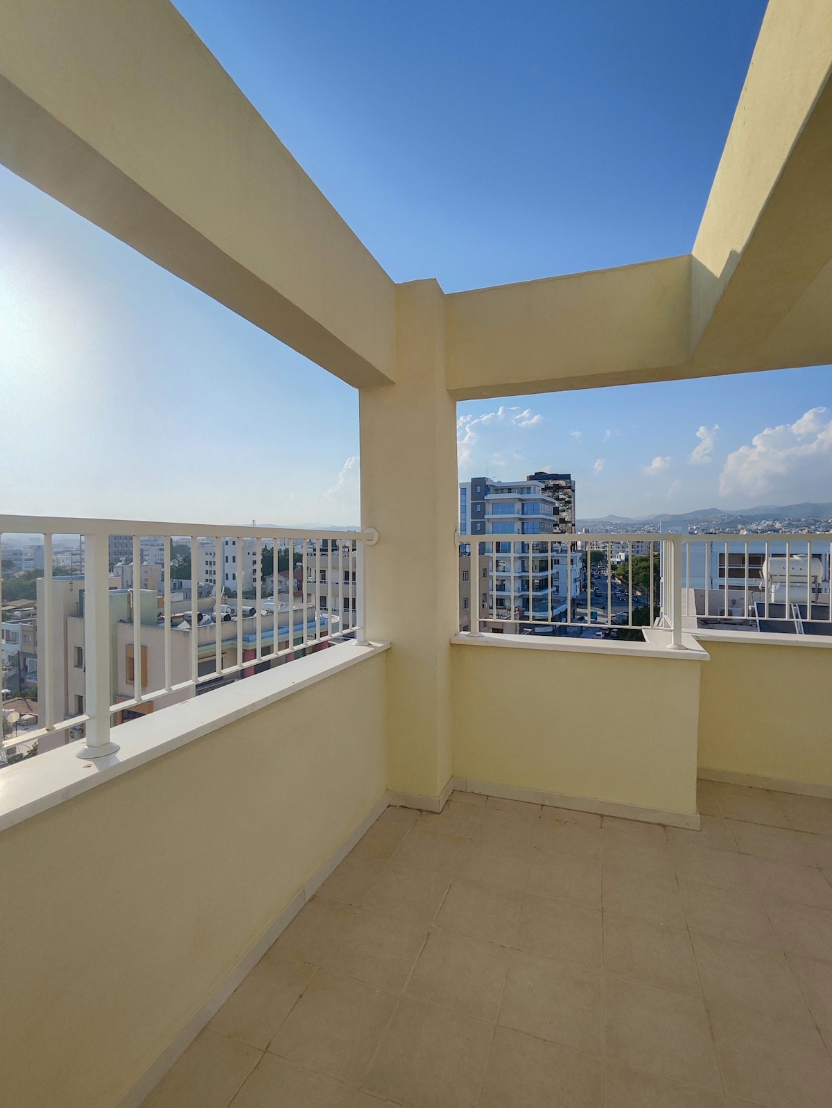 A spacious private terrace is adorned with a white safety railing, providing unobstructed views of the city skyline and surrounding mountains. Sunlight illuminates the pale walls and tiled floor, creating an inviting outdoor space for relaxation.