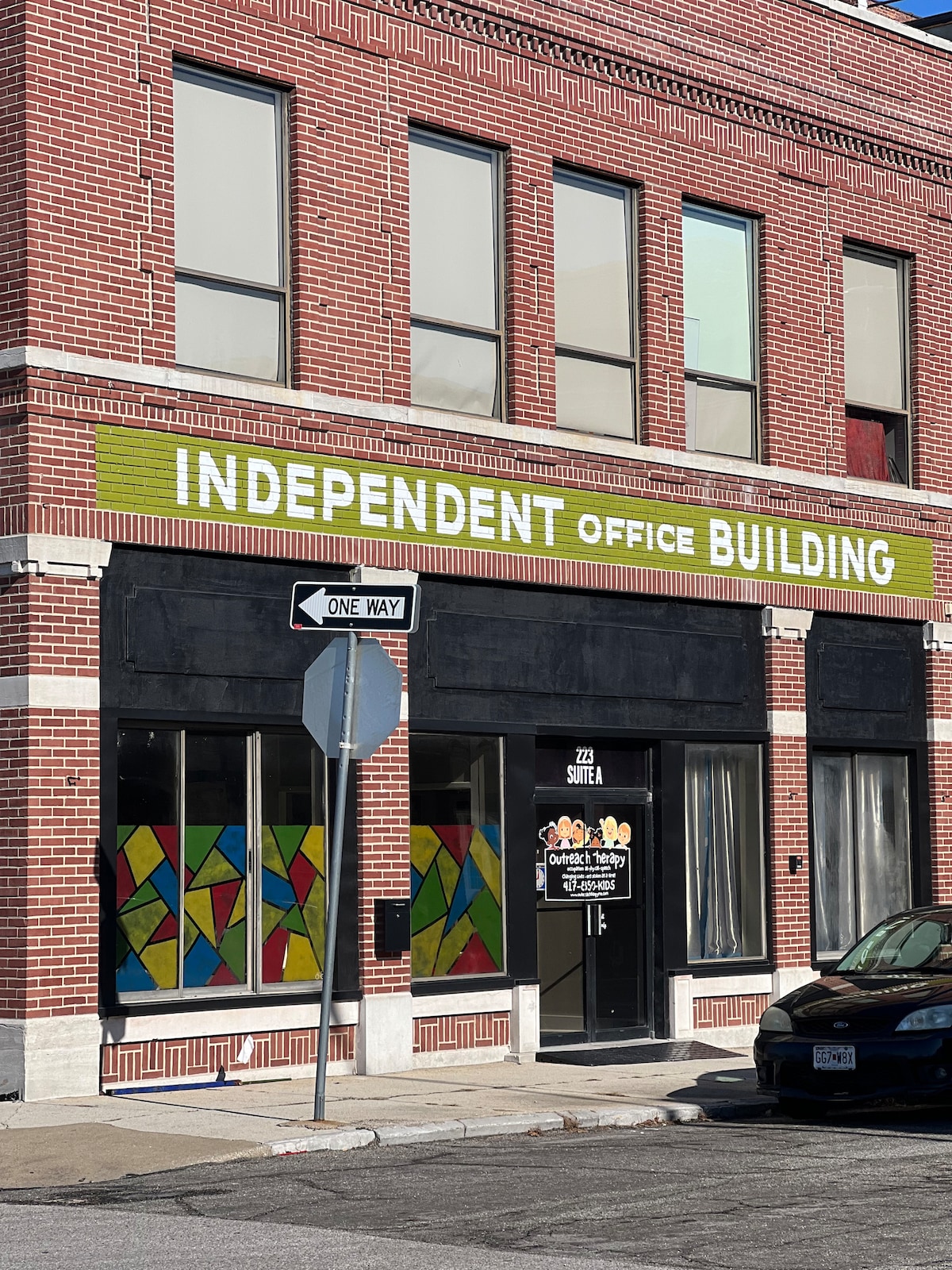 The image showcases a historic brick building with large windows and a vibrant green sign labeled 'INDEPENDENT OFFICE BUILDING.' Colorful stained glass panels adorn the lower section, providing a striking contrast against the black storefront. A one-way street sign is visible in the foreground.