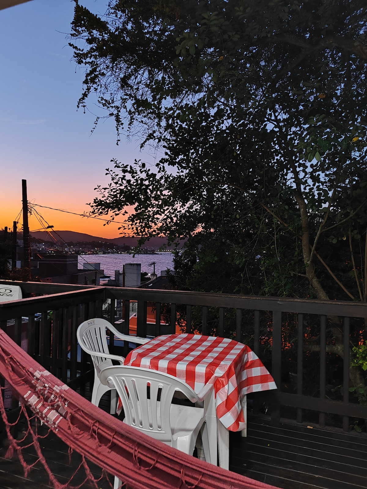 A deck area offers a view of a sunset over the water, framed by greenery. A red and white checkered tablecloth covers a table, accompanied by two white chairs. A hammock in the foreground invites relaxation in the evening ambiance.