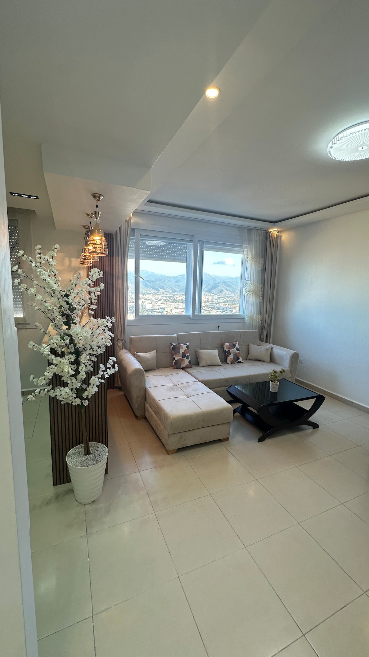 A modern living area features a light-colored sectional sofa against a wall adorned with floor-to-ceiling windows, offering expansive views of the surroundings. A small coffee table sits in front, accompanied by decorative items, while a tall vase with artificial flowers adds a touch of elegance.