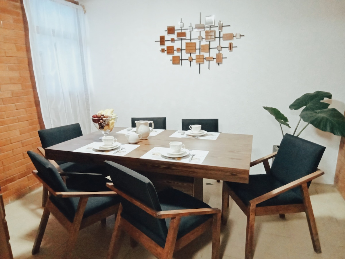 A dining area features a wooden table surrounded by six dark chairs. A modern wall art piece is displayed above the table, and a small vase with fruit is present. Natural light is allowed in through a nearby curtain-covered window.