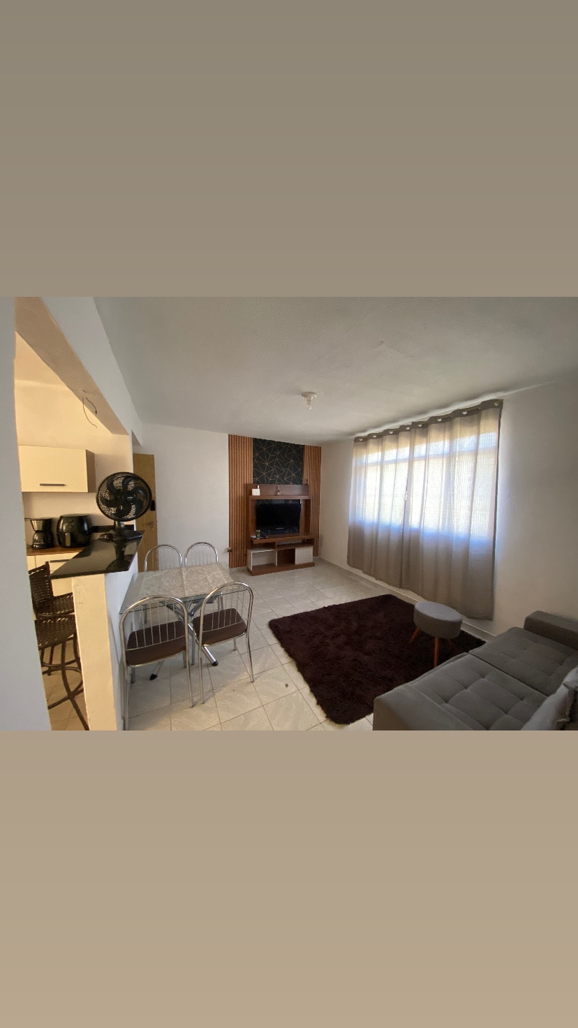 A spacious living area is shown, featuring a comfortable gray sofa, a round coffee table, and a small dining set with metal chairs. Natural light filters through large, sheer curtains, while a wooden television unit is positioned against the wall, adding functionality to the space.