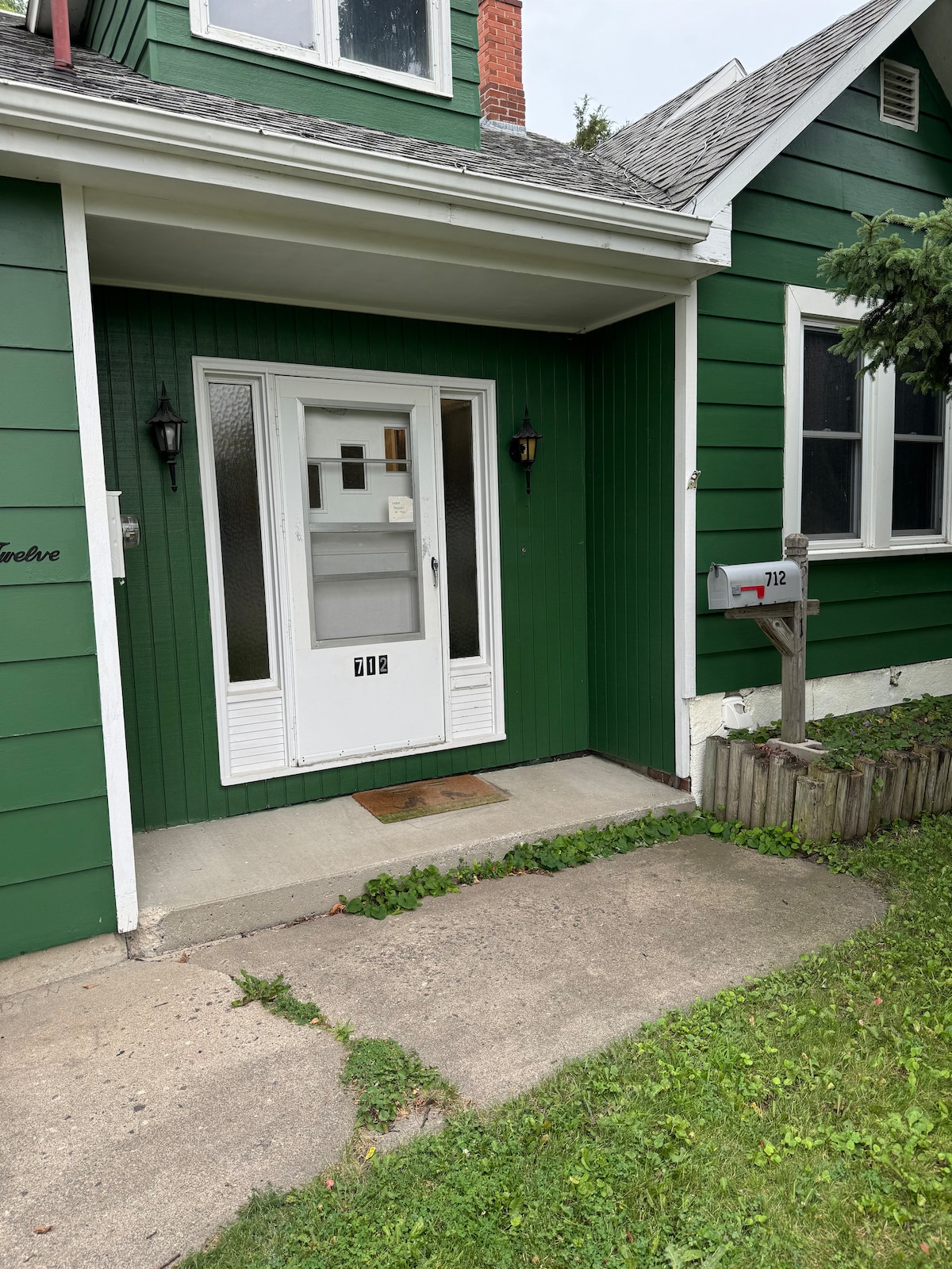 An entrance to the little house is framed by green siding and a well-maintained lawn. A white front door with glass panels is centered, flanked by two wall-mounted lights. The mailbox stands to the right, beside a narrow flower bed.