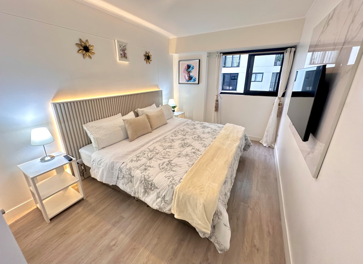 A spacious bedroom features a large bed with neutral-toned bedding and decorative pillows. Accent lights are positioned behind the headboard, providing soft illumination. A nightstand with a lamp sits beside the bed, and a wall-mounted television is installed across from the sleeping area.