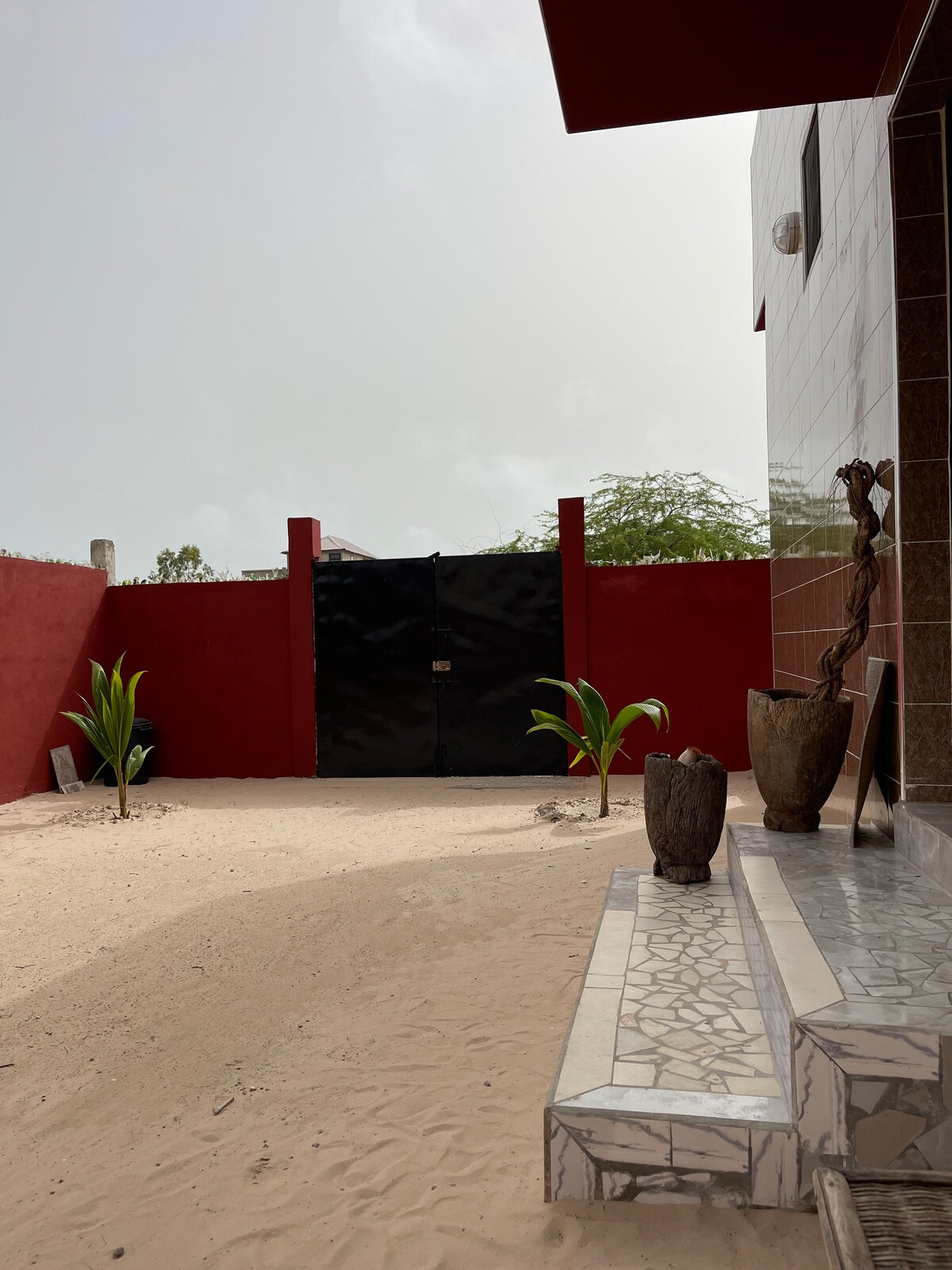 An entrance is framed by sandy ground with minimalist landscaping. A pair of small plants are positioned at the base of a tiled staircase leading to a black gate. Textured walls in red and light brown provide a bold backdrop.
