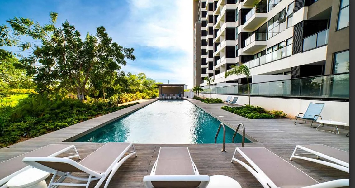 The image features a modern swimming pool surrounded by a wooden deck and lounge chairs. Lush greenery is visible on the sides, providing a natural contrast to the stylish building in the background. Clear blue skies and sunlight enhance the inviting atmosphere of the space.