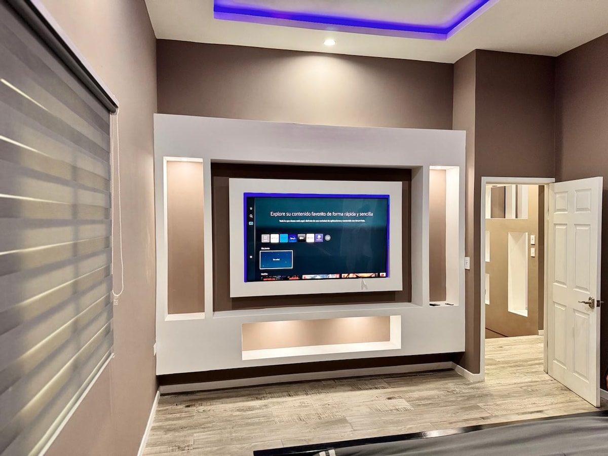 A modern living area features a sleek entertainment unit with a large flat-screen television mounted on a white wall. Soft purple lighting runs along the ceiling, while a light-tone floor enhances the contemporary design. A closed window with dual-layer blinds is visible on the left.
