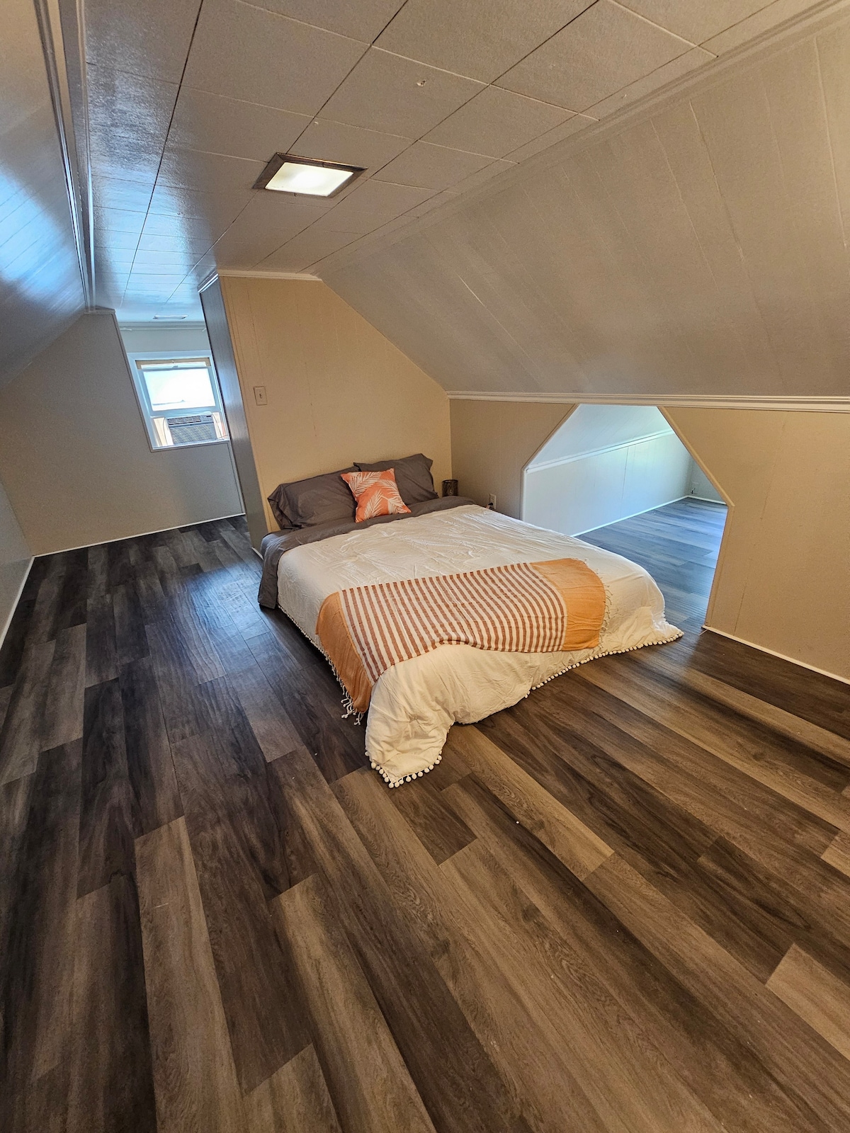 A cozy attic bedroom features a spacious bed adorned with a striped blanket. Natural light enters through a nearby window, illuminating the wood-like flooring. Unique architectural details are highlighted by angled ceilings and a simple color palette, creating a serene atmosphere.
