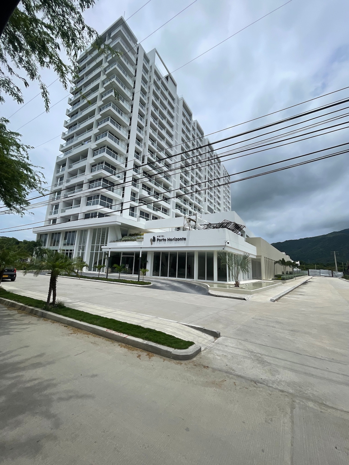 A modern high-rise building stands prominently against a cloudy sky. The structure features multiple layers of balconies and large windows. The entrance is framed by neatly landscaped green areas and a spacious parking lot, highlighting the contemporary architectural design.
