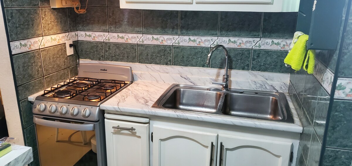 A compact kitchen is shown, featuring a gas stove with four burners and an oven. A double sink sits alongside a marble countertop, complemented by green tiled walls with decorative trim. Under-cabinet lighting provides additional illumination, enhancing the workspace.