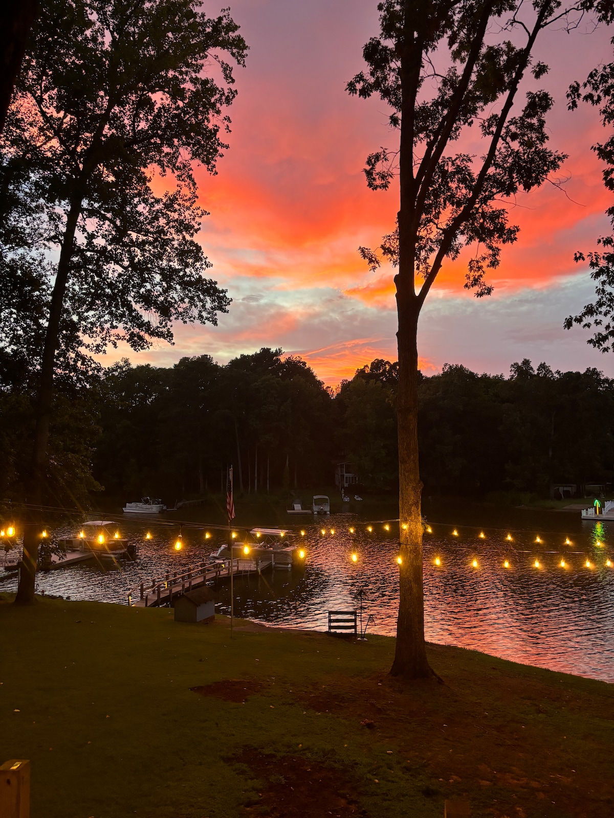 A serene evening scene along the lake is captured, featuring a vibrant sunset sky with hues of orange and pink. Soft twinkling lights are strung along the dock, reflecting on the calm water, while trees frame the peaceful view.