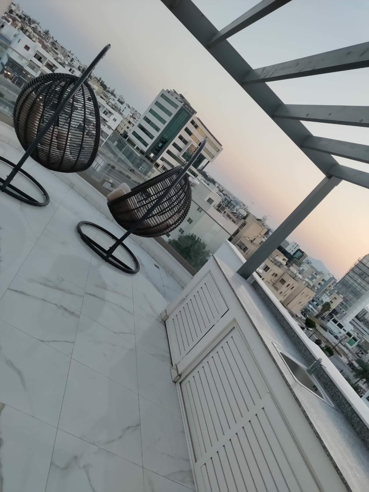Two hanging chairs are positioned on a spacious shared rooftop, overlooking the cityscape of Larnaca. The terrace features a marble surface and a white railing, with a backdrop of modern buildings and a soft gradient sky during sunset.