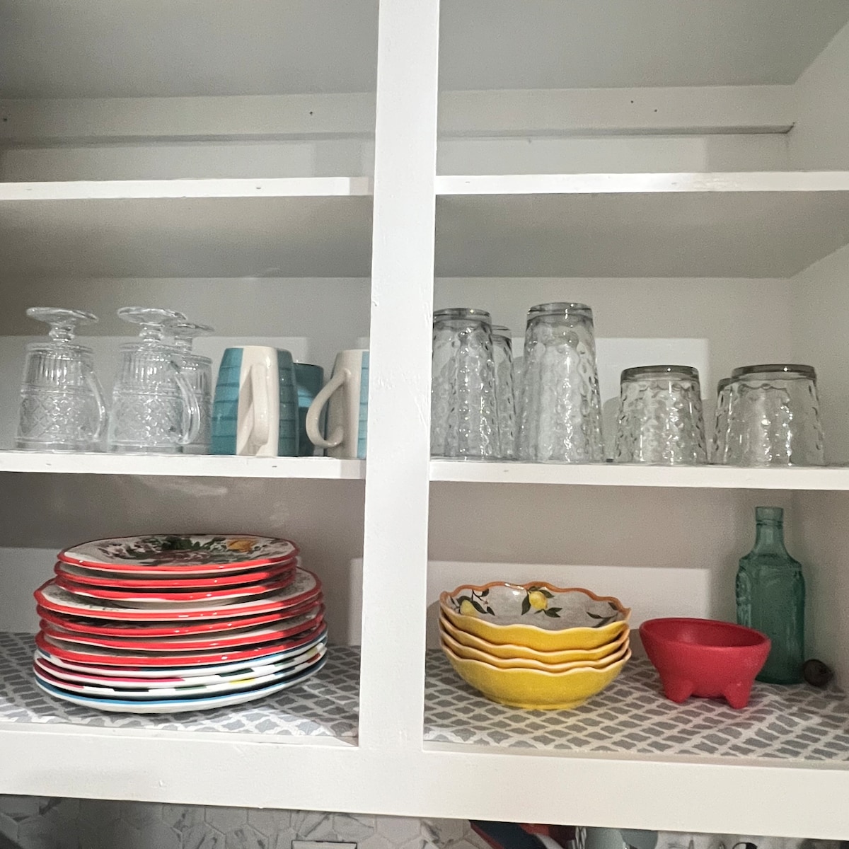 A neatly organized kitchen cabinet features colorful plates stacked in a circular arrangement. Glassware is displayed on the upper shelf, with various glasses and pitchers visible. A set of decorative bowls in vibrant colors is also present, alongside a small green glass bottle.