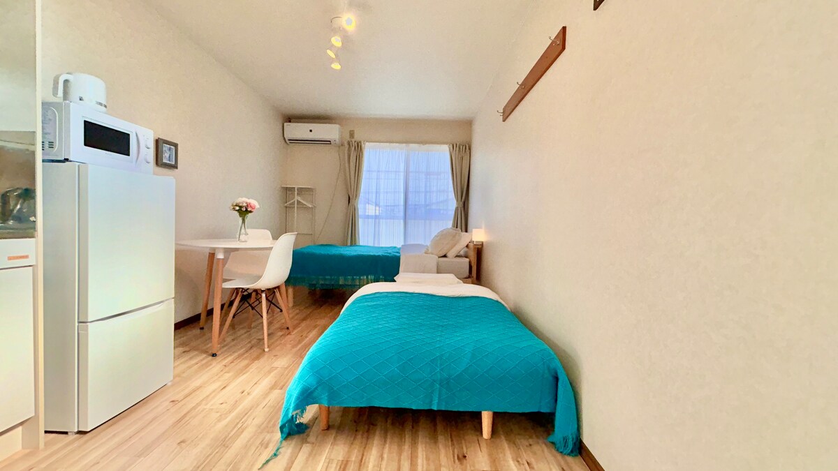 A bright, open studio space is displayed with soft lighting. Two beds are neatly arranged, one adorned with a turquoise coverlet. A dining table with modern chairs is positioned nearby, alongside a refrigerator and a small kitchenette. Large windows allow natural light to fill the room.