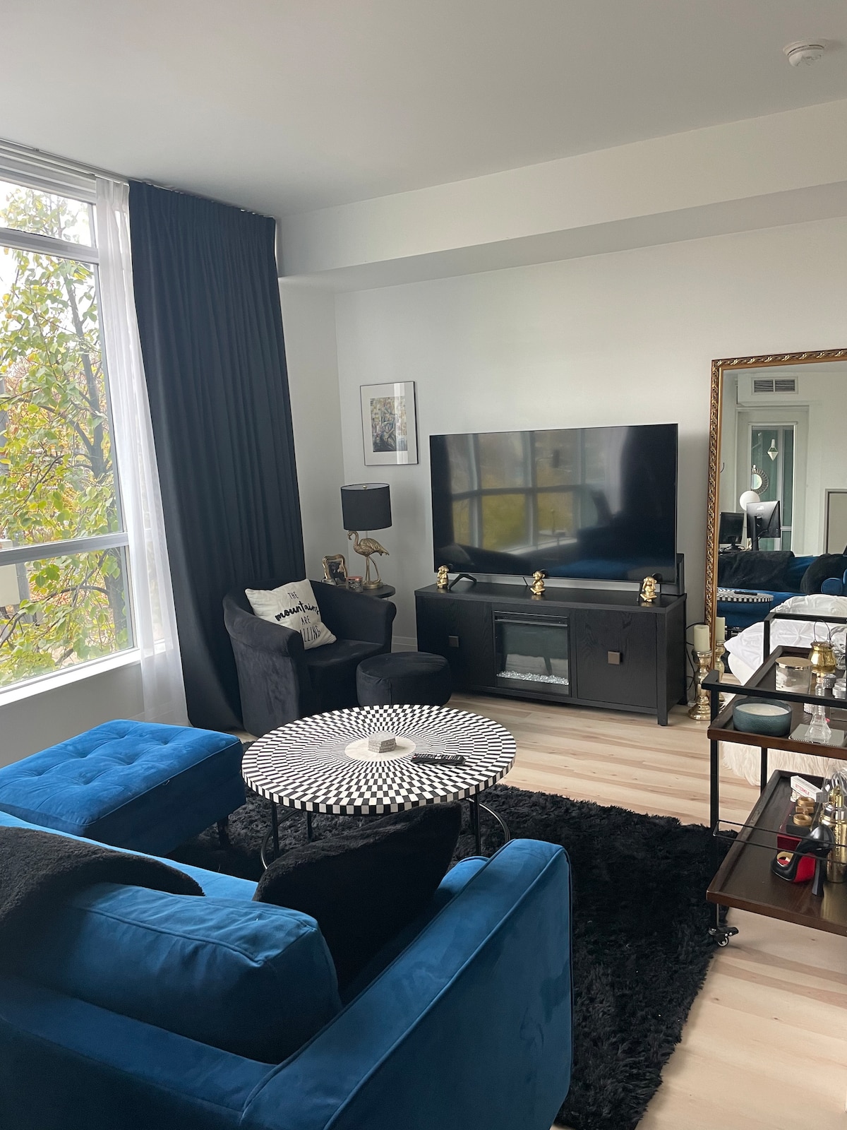 A bright living area features large windows showcasing natural light, with dark drapes framing the view. A blue velvet sofa and armchair are arranged around a patterned coffee table, while a sleek media console with a television is positioned against the wall.
