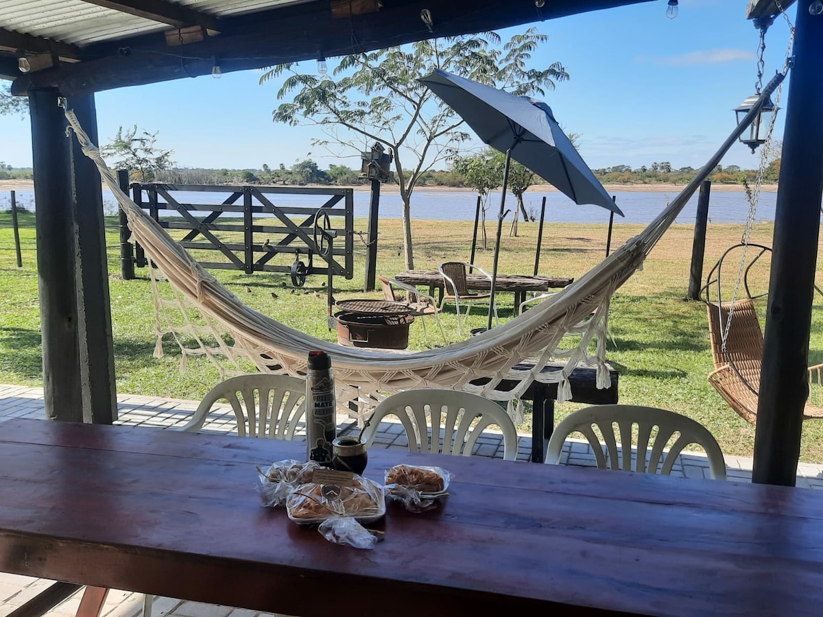 A cozy outdoor space features a rustic table at the forefront, accompanied by white plastic chairs. A hammock hangs nearby, inviting relaxation. A large umbrella offers shade, while a scenic view of the river and grassy area completes the serene atmosphere.