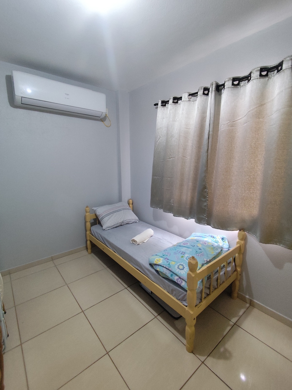 A snug room features a single bed with a light blue bedspread and a rolled towel placed neatly on top. Soft light filters through partially closed, beige curtains, and a wall-mounted air conditioning unit is visible. The floor is tiled for easy maintenance.