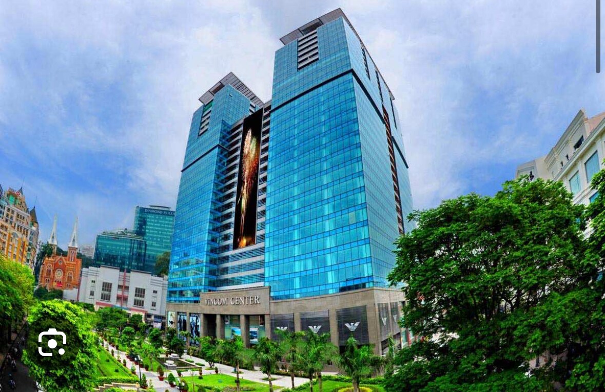 A large, modern building features a façade of reflective glass with a central vertical display. Surrounding greenery includes lush trees and meticulously maintained gardens, contributing to the inviting landscape. The structure is situated within a vibrant urban setting, with city landmarks visible in the background.
