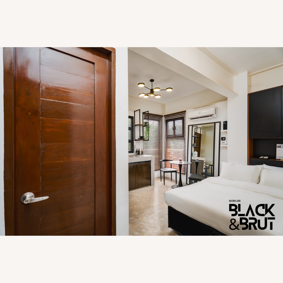 The entrance to the studio is visible, showcasing a wooden door leading into a modern space. Natural light streams through window blinds, illuminating the cozy sleeping area with a spacious bed. A sleek vanity and minimalist decor are present, enhancing the contemporary design.