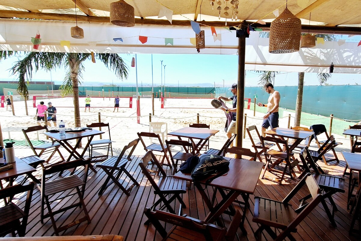 An outdoor dining area features multiple wooden tables and folding chairs under a shaded structure. The sandy beach tennis court is visible in the background, with vibrant colors and recreational activity occurring nearby. Decorative elements, such as hanging lanterns and colorful banners, enhance the setting.