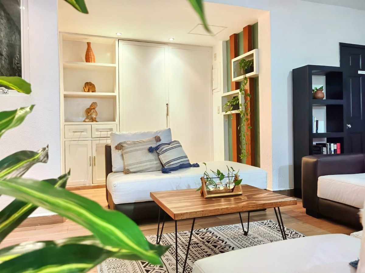 A bright living area features contemporary furniture with a neutral color palette. A wooden coffee table sits in the center, adorned with a small plant. Soft seating is complemented by decorative cushions, and built-in shelves display decorative items and greenery.