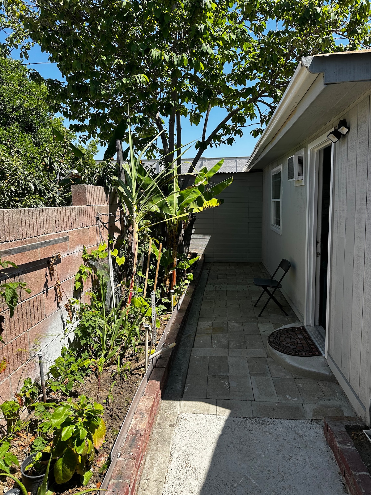 A narrow pathway is visible beside the building, bordered by a modest garden featuring various plants and trees. Sunlight filters through the greenery, highlighting the textured stone paving. A small chair is set against the wall, providing a quiet spot to enjoy the outdoor space.