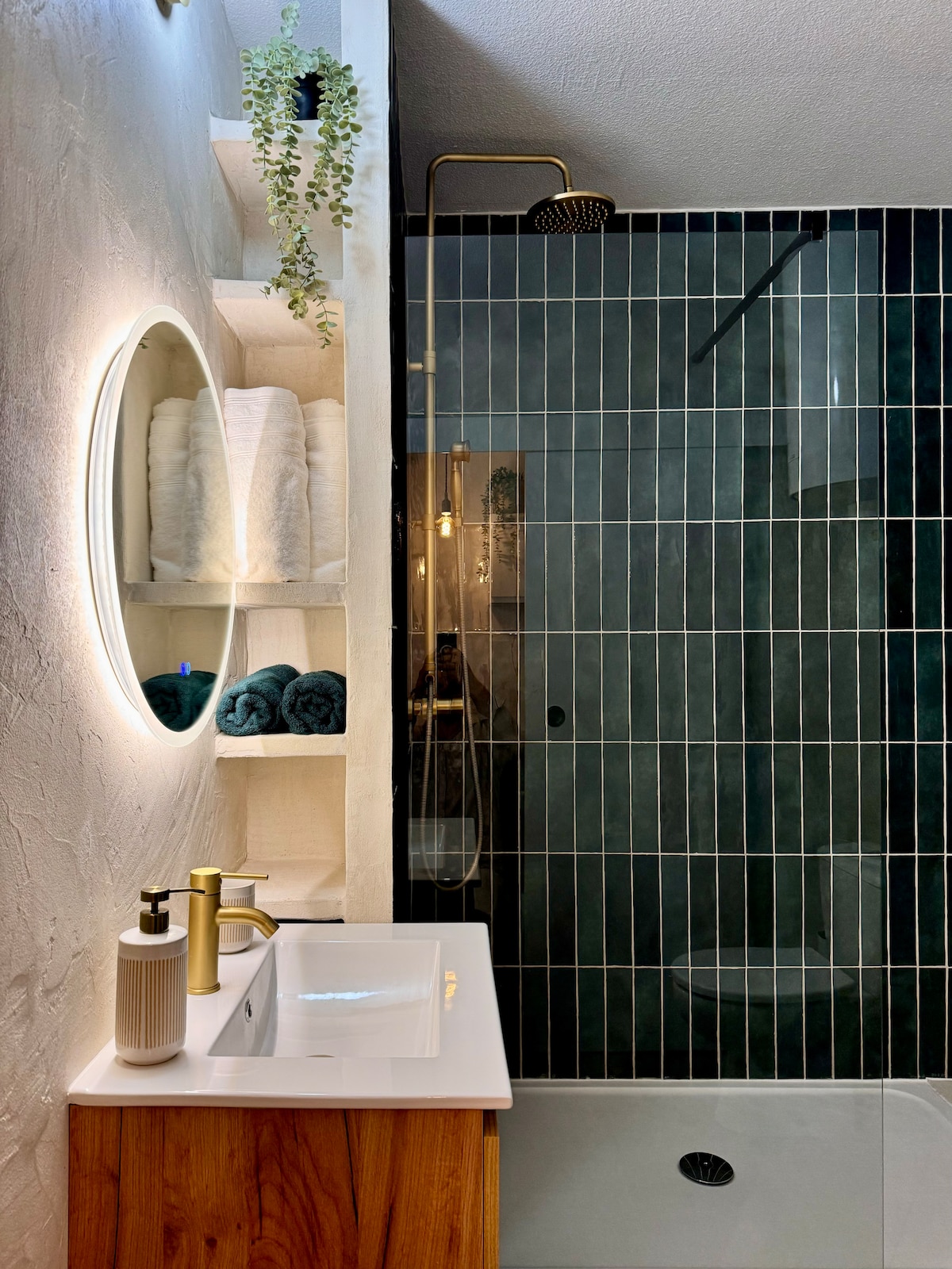 A modern bathroom features a walk-in shower with dark tile and a rainfall showerhead. A round mirror is backlit above a wooden vanity with a white countertop. Shelves display neatly arranged towels and decorative greenery, adding a touch of freshness.