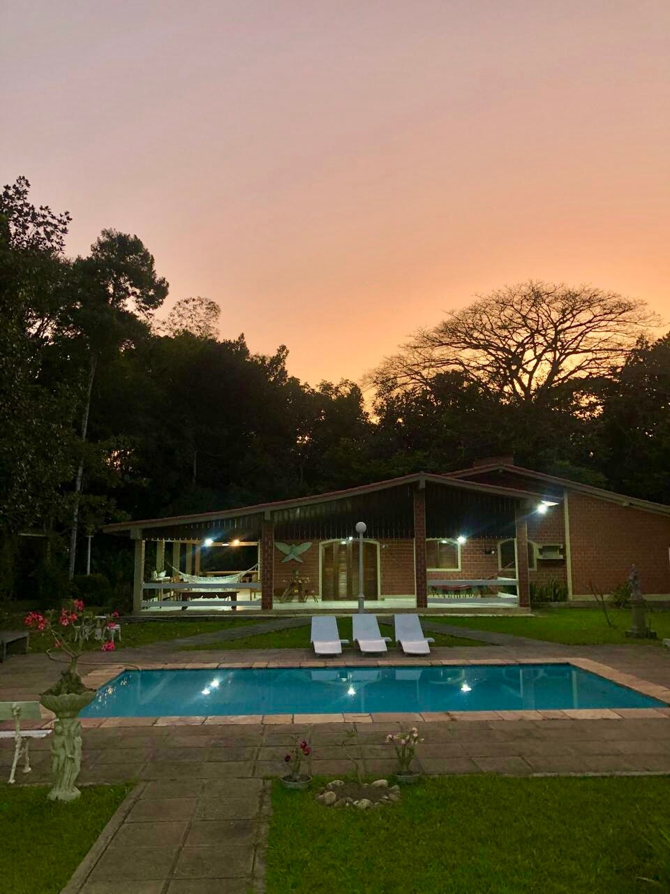 A rustic house is nestled among trees, featuring large windows that provide views of the surrounding nature. In the foreground, a sparkling pool is flanked by two lounge chairs, creating a serene outdoor relaxation area. The sky displays warm hues at dusk, enhancing the tranquil setting.