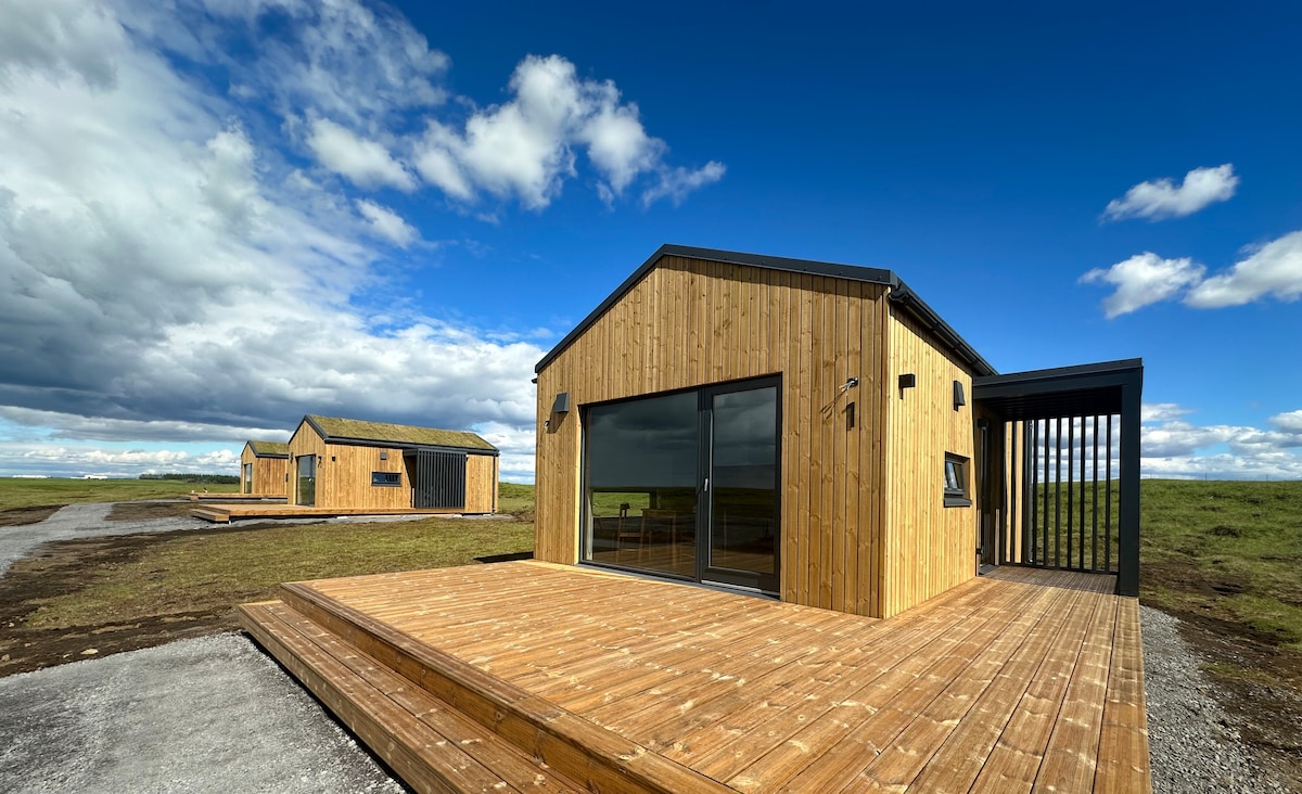 Two small wooden cottages are situated on a gravel path, surrounded by expansive green fields. Each cottage features large glass doors that reflect the sky, inviting natural light indoors. The exterior showcases a contemporary design with wooden siding and a flat roof.
